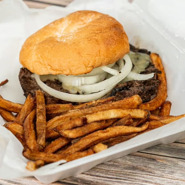 Cheeseburger with onions and a side of fries in a white takeout container on a wooden table.