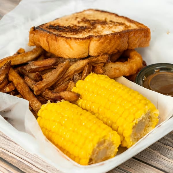 Grilled sandwich with a side of fries, onion rings, two corn on the cob, and a dipping sauce.