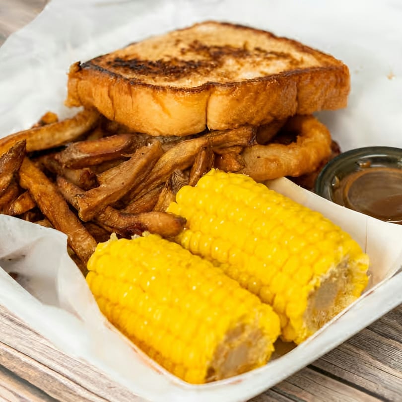 Grilled sandwich with a side of fries, onion rings, two corn on the cob, and a dipping sauce.