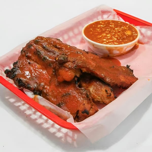 Barbecue ribs with sauce in a red basket lined with white paper, alongside a small cup of baked beans.